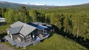 Uma casa moderna com um deck, rodeada por uma floresta e montanhas ao fundo.