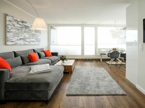 A modern living room with a grey sofa, a coffee table, and a rug. There is a dining area with a table and chairs in the background. - Holiday Apartment With 1 Bedroom (Sylt)