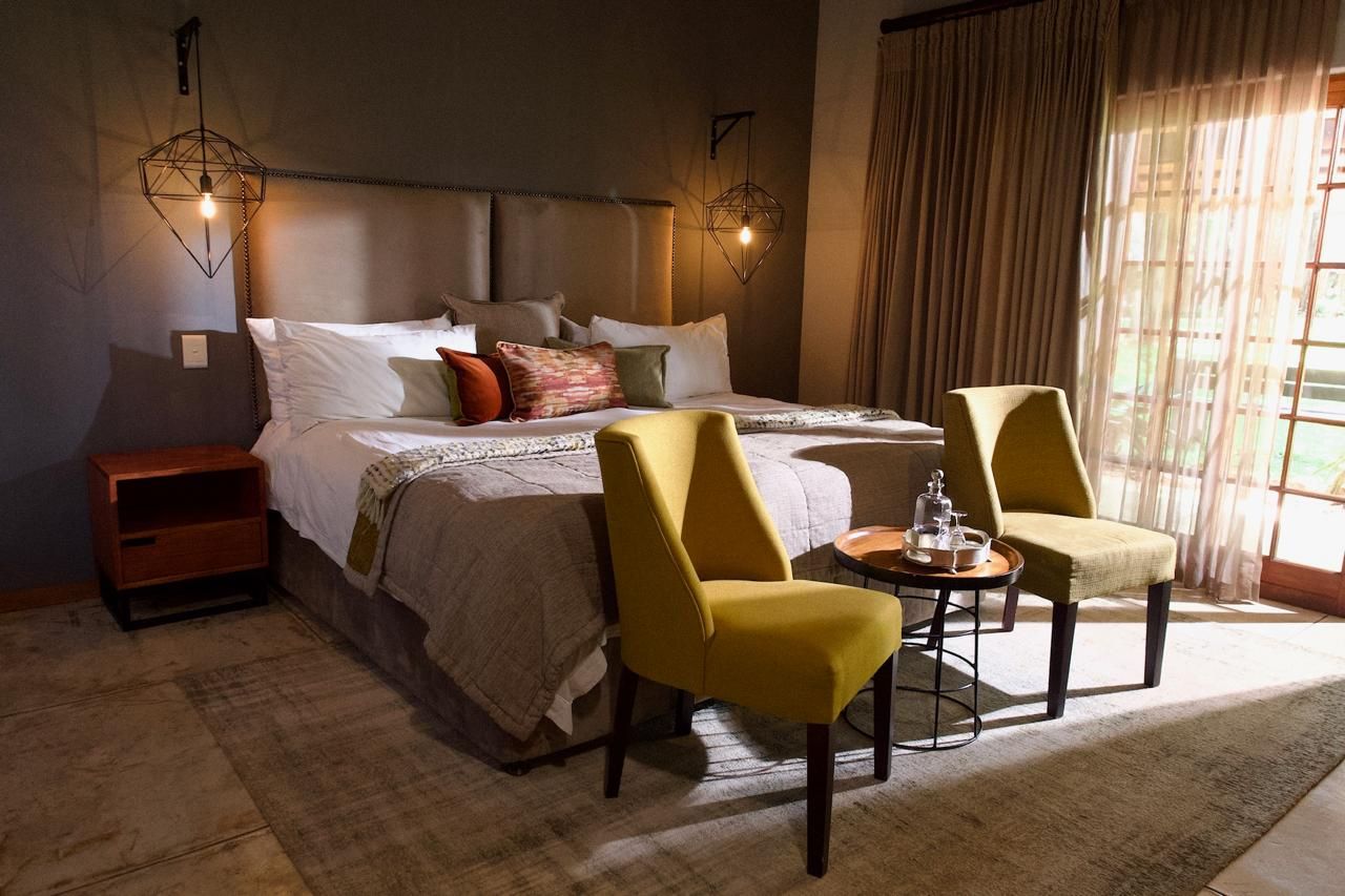 Quarto de hotel com uma cama grande, duas cadeiras, uma mesinha e uma mesinha de cabeceira.