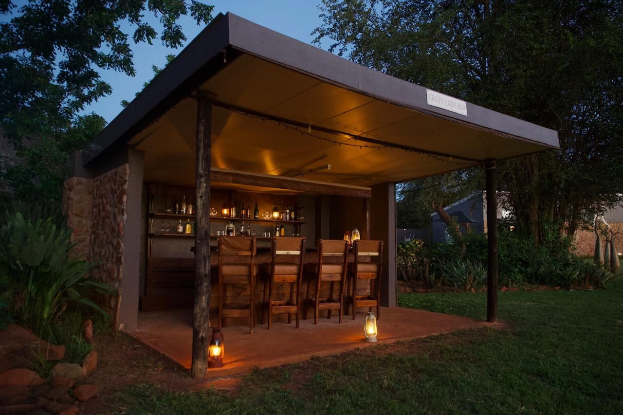 An outdoor bar area with wooden chairs and tables, surrounded by stone walls and greenery.