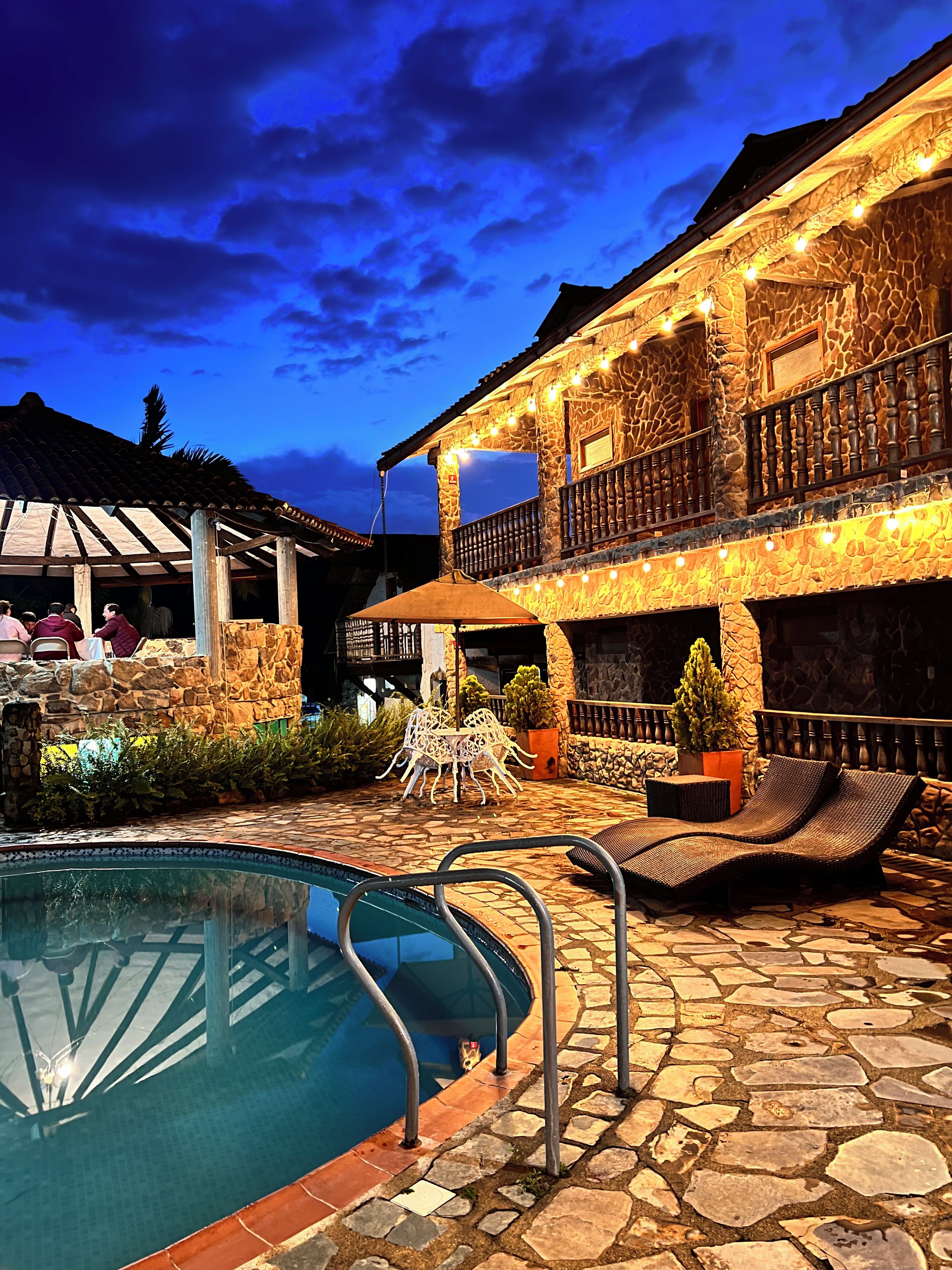 Zona de piscina con suelo de piedra, sillones de descanso y un edificio con paredes y balcones de piedra.