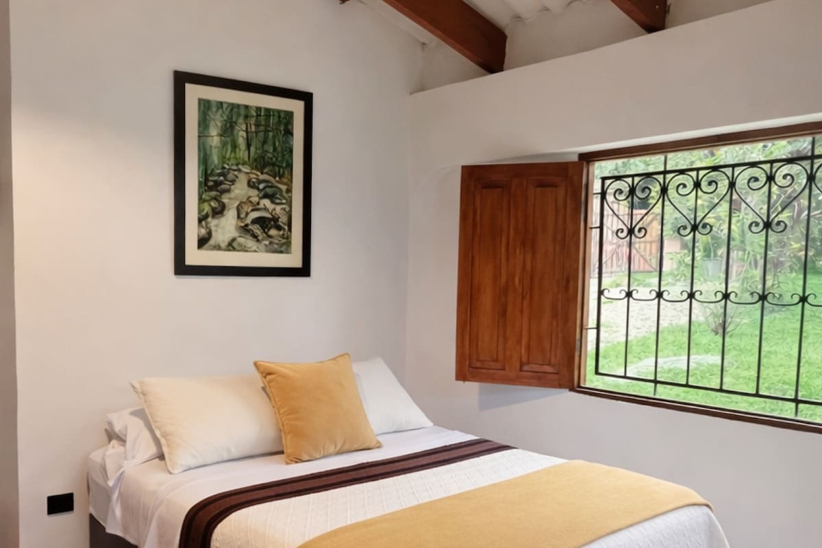 A bedroom with a bed, a window with a view, a ceiling fan, and a framed picture on the wall.