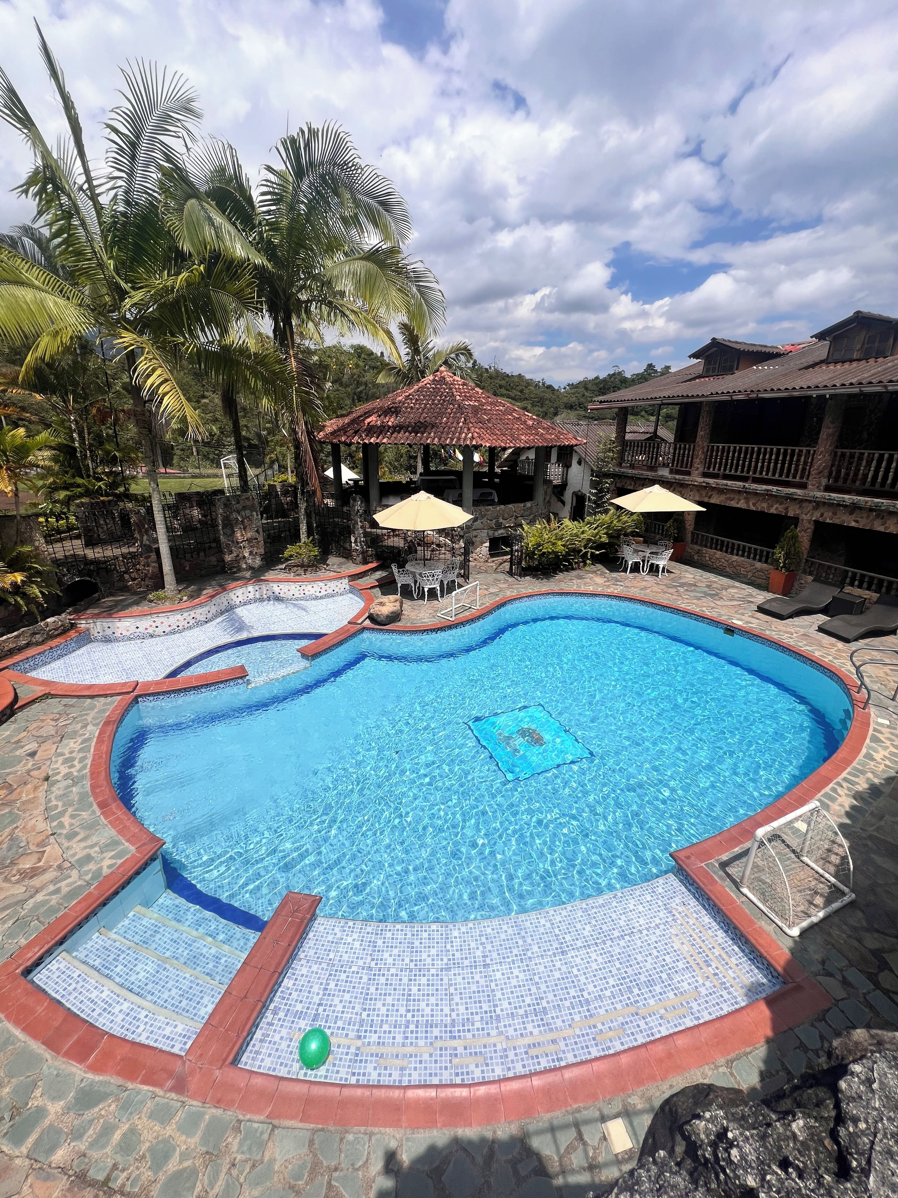 A swimming pool with a tiled deck, surrounded by palm trees and a gazebo.