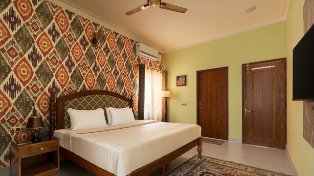 A bedroom with a bed, nightstand, lamp, patterned wall, ceiling fan, and two doors. - Casa Albela (Bengaluru)