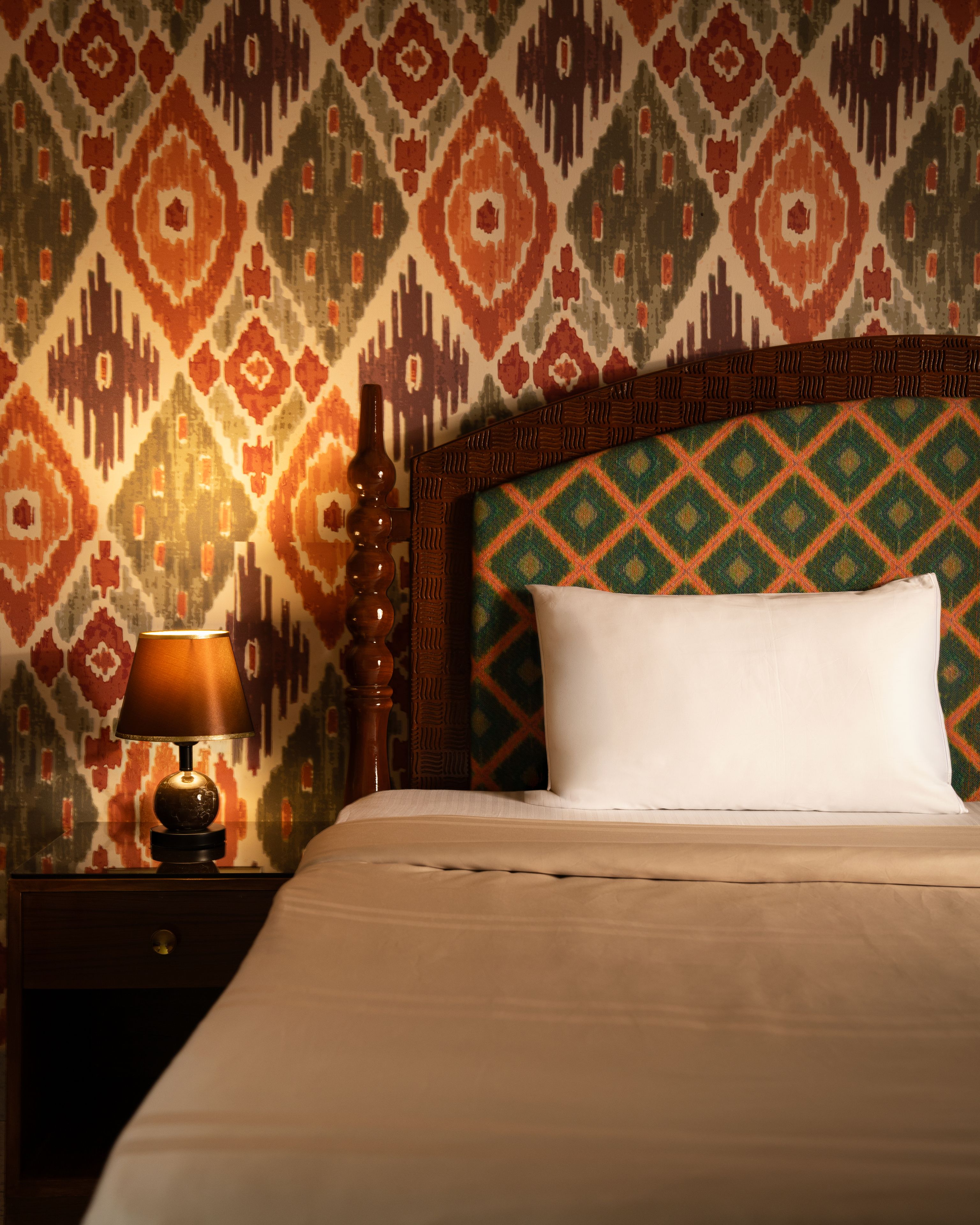 Een hotelkamer met een behang, een houten hoofdbord, een wit kussen en een nachtkastje met een lamp.