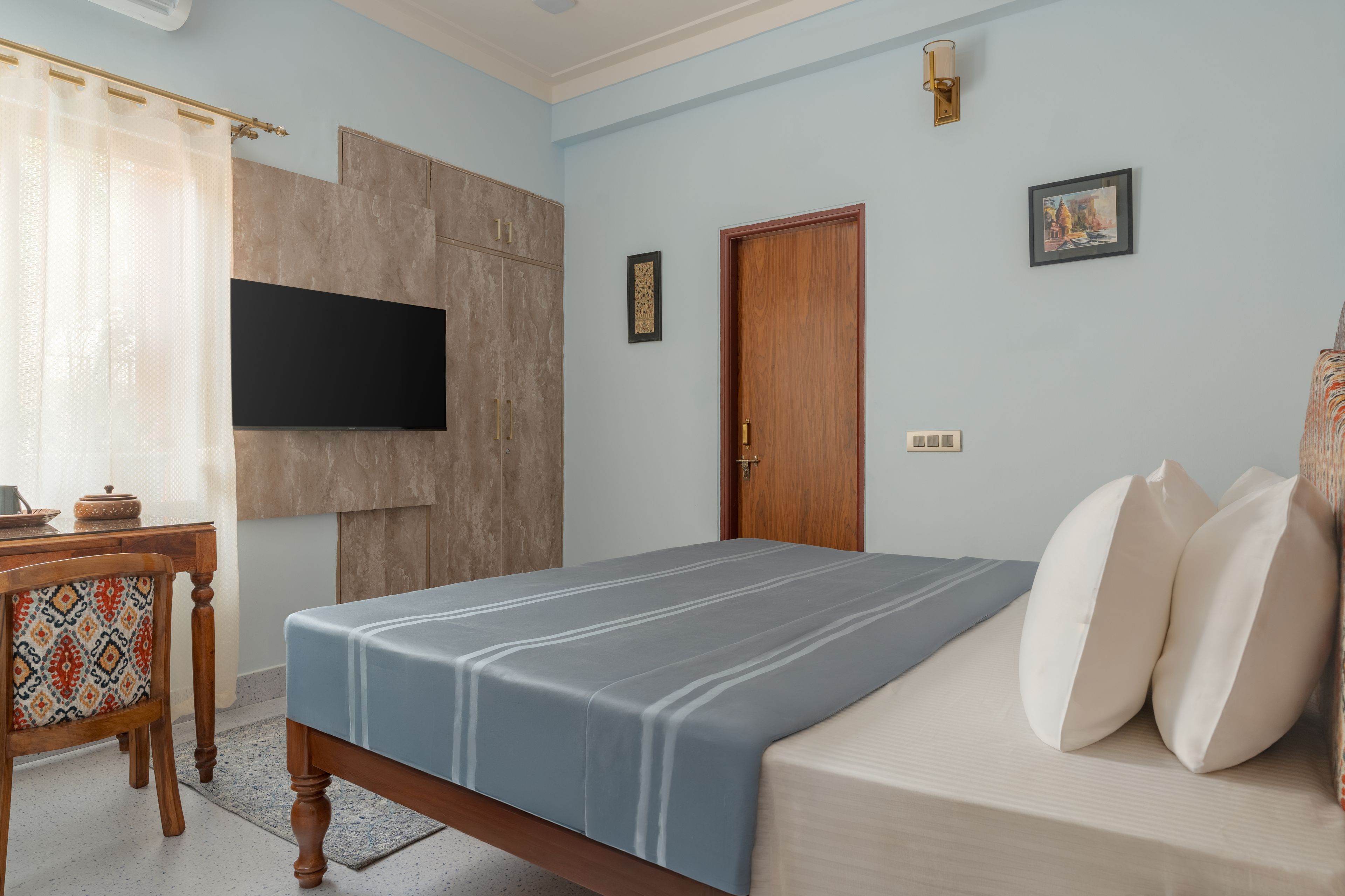 A bedroom with a bed, a desk, a television mounted on the wall, and a door.