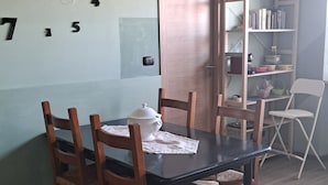 Een eetruimte met een tafel gedekt voor zes personen, omringd door houten stoelen. Op tafel staat een witte theepot, en er is een houten deur in de achtergrond te zien.