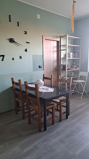 Un'area pranzo con un tavolo apparecchiato per sei persone, circondato da sedie di legno. Sulla tavola c'è un teiera bianca, e sullo sfondo si intravede una porta in legno.