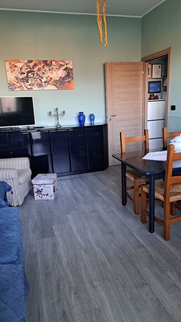 Un soggiorno con divano, televisione, tavolo da pranzo con sedie, angolo cottura con pensili e un quadro appeso al muro.