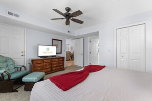 Ein Schlafzimmer mit einem Bett, einem Fernseher, einem Schminktisch, einem Sessel und einem Deckenventilator.