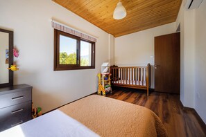 A bedroom with a wooden ceiling, a crib, a bed with a beige quilt, a dresser, and a window with a view of greenery. - Troodos View Garden House - Trimiklini (Trimiklini)
