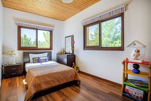 A bedroom with a wooden ceiling, a bed with a patterned bedspread, a dresser, a nightstand with a lamp, and a bookshelf with board games. - Troodos View Garden House - Trimiklini (Trimiklini)