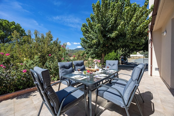 Eine Außenpatio mit einem für das Essen gedeckten Tisch, umgeben von Stühlen, und ein großer Baum spendet Schatten.