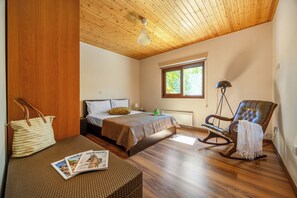 Ein Schlafzimmer mit Holzbalkendecke, einem Bett mit Kopfteil, einem Schaukelstuhl, einem Fenster und einer Zeitschrift auf einem Nachttisch.