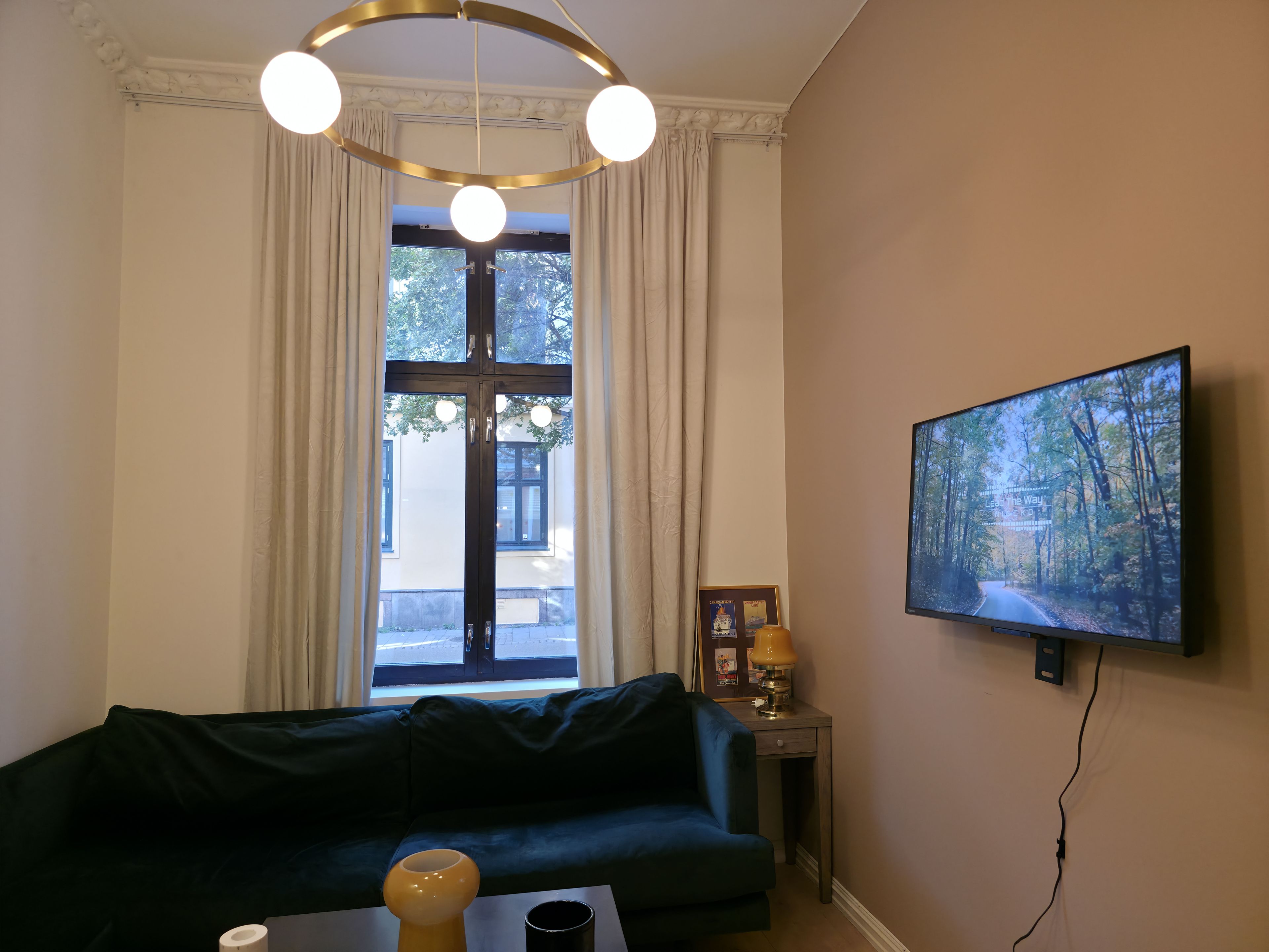 A room with a dark green sofa, a small table with a lamp, a framed picture, and a television displaying a forest scene.