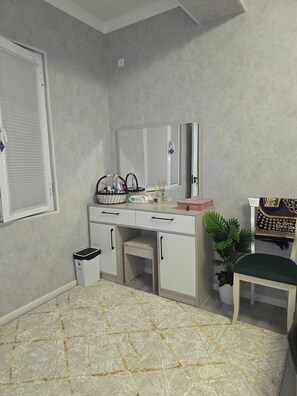 A bedroom with a bed, a desk, a chair, a mirror, a window with blinds, a trash bin, and a potted plant.