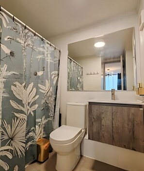 Et badeværelse med bruseforhæng med bladmotiv, et hvidt toilet, et træbord med spejl og et vindue med udsigt.