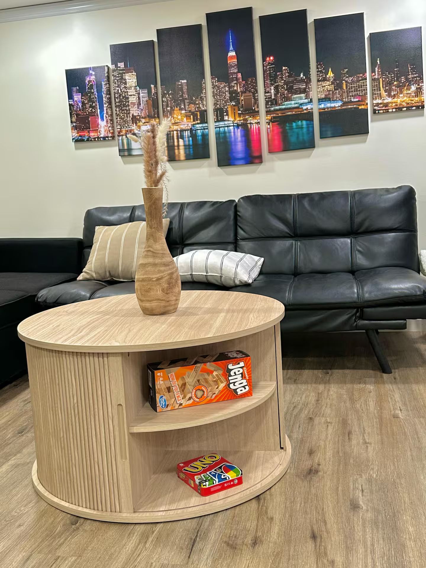 Una sala de estar moderna con un sofá de cuero negro, una mesa de centro de madera con un jarrón y una caja de juegos, y una vista de la ciudad en la pared.