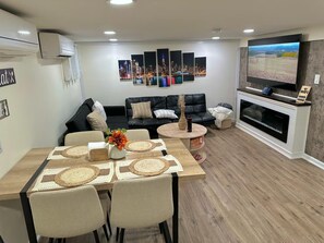 A modern living room with a dining area, a black leather sofa, a flat-screen TV, and a fireplace.