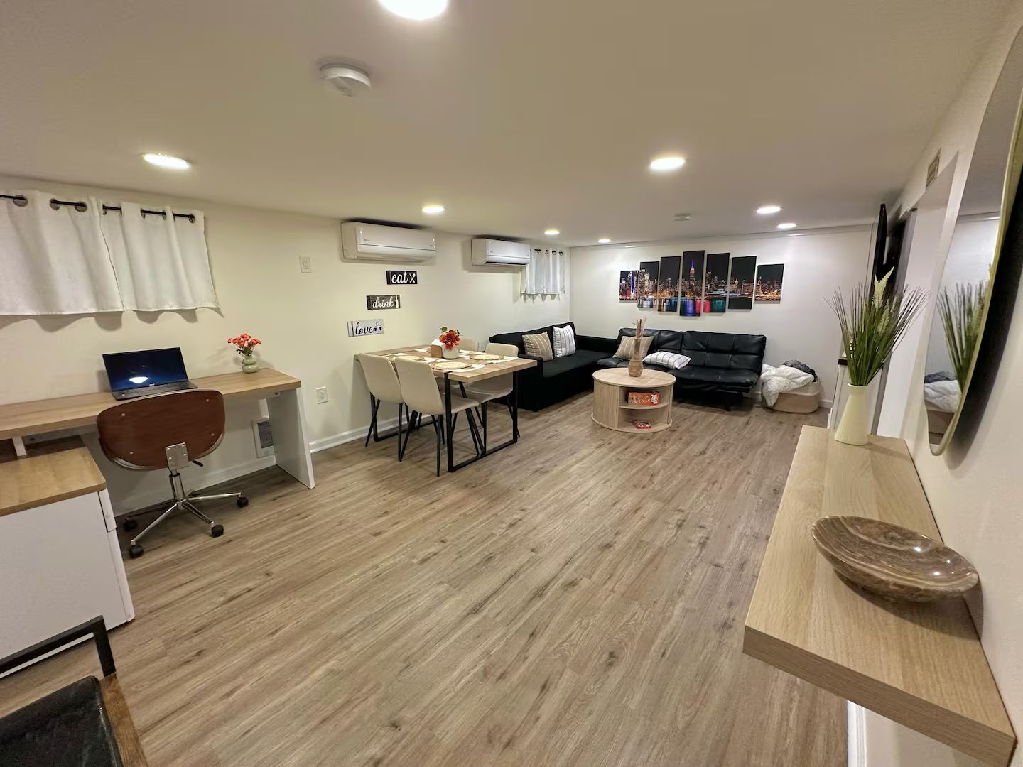 Um espaço de estar moderno com área de jantar, sofá de couro preto, mesa de centro de madeira e um laptop em uma escrivaninha.
