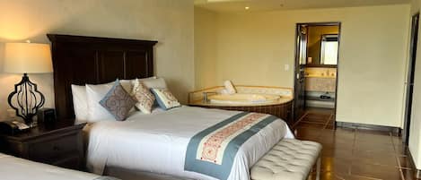 A hotel room with two beds, a bench, a nightstand, a lamp, a bathroom with a sink and mirror, and a tiled floor.