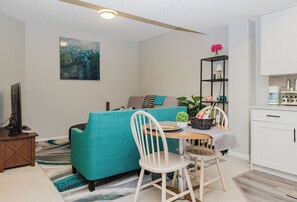 Ein modernes Wohnzimmer mit einem türkisfarbenen Sofa, einem weißen Sessel, einem kleinen Esstisch und einer Küchenzeile mit weißen Schränken und einem schwarzen Regal.