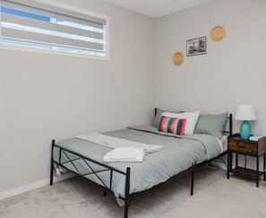 A bedroom with a bed, a nightstand, a lamp, a window with blinds, and two wall decorations. - Modern 1-Bedroom, 1- Bathroom Private Basement Suite in Windermere (Edmonton)