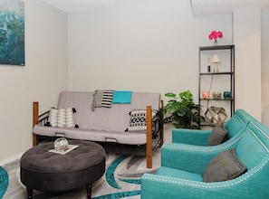 Ein Wohnzimmer mit einer Couch, einem Hocker und einem türkisfarbenen Sessel, einem Bücherregal mit Dekoration und einer großen Pflanze.