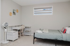 Ein Schlafzimmer mit Bett, Schreibtisch mit Stuhl, Standventilator und einem Fenster mit Jalousien.