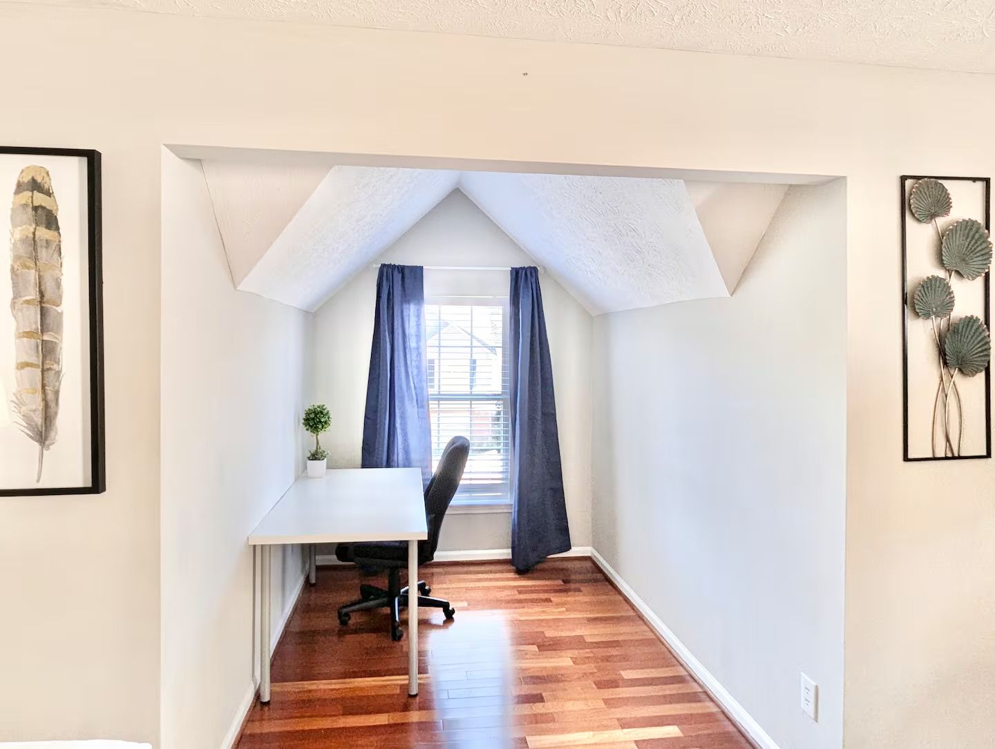 Un pequeño cuarto ático triangular con escritorio, silla y ventana con cortinas.