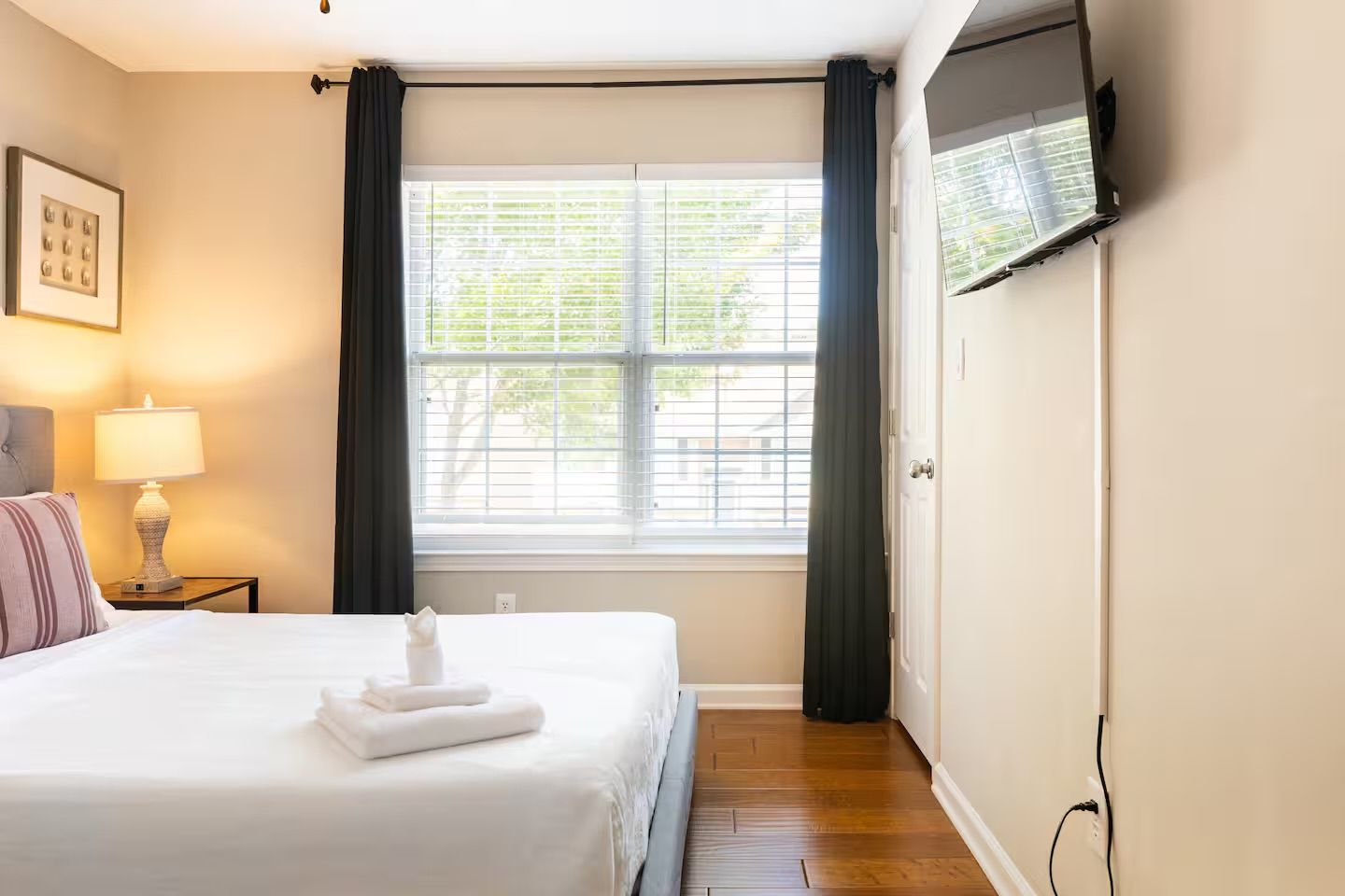 Habitación de hotel con cama, lámpara de noche, ventana con cortinas y televisor montado en la pared.