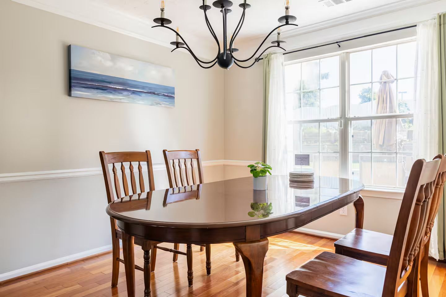 A dining room with a round table, wooden chairs, a chandelier, a painting, and a window with curtains.
