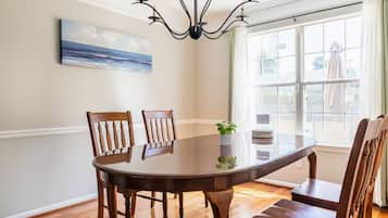 A dining room with a round table, wooden chairs, a chandelier, a painting, and a window with curtains.