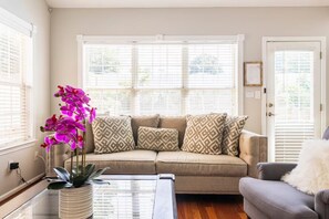 Una sala de estar con un sofá color beige, cojines estampados, una mesa de centro de vidrio y una planta en maceta con flores moradas.