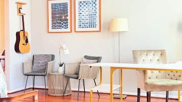 Una sala de estar con una guitarra de madera, dos cuadros abstractos enmarcados, una lámpara y un comedor con una mesa y sillas blancas.