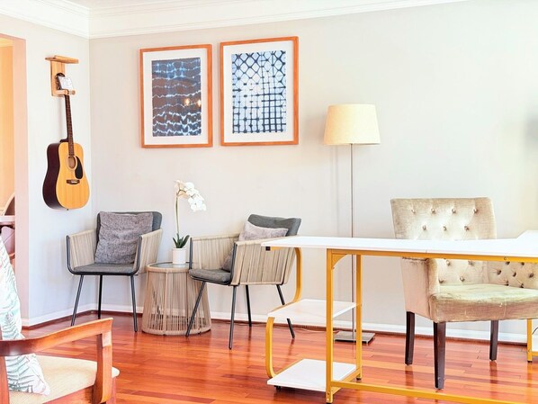A living room with a wooden guitar, two framed abstract paintings, a lamp, and a dining area with a white table and chairs.
