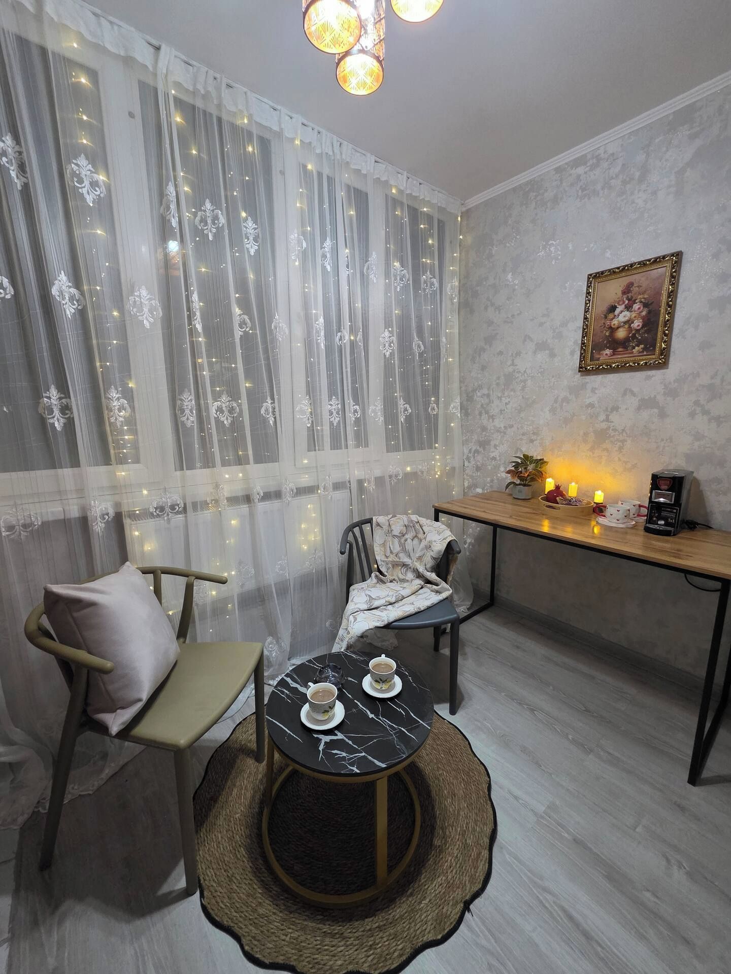 A cozy room with a dining table, chairs, a small round table with cups, a decorative wall piece, and a window with sheer curtains.