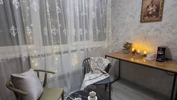 A cozy room with a dining table, chairs, a small round table with cups, a decorative wall piece, and a window with sheer curtains.