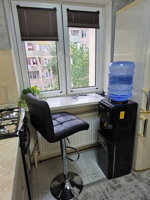 Una cocina con estufa a gas, un taburete negro, dispensador de agua y vistas de edificios a través del ventanal.