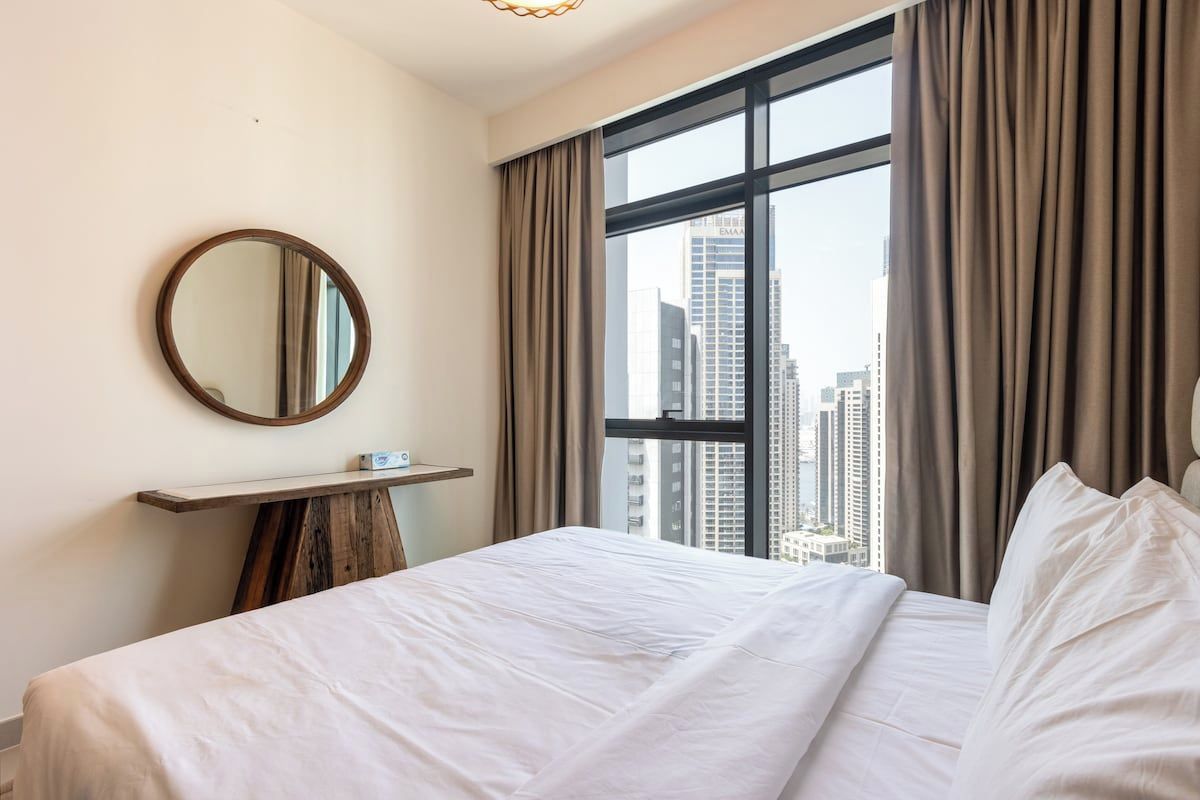 Ein Hotelzimmer mit großem Fenster, einem runden Spiegel, einem Nachttisch und einem Bett mit weißer Bettwäsche.