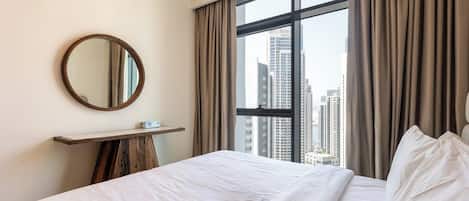 Habitación de hotel con un ventanal grande, un espejo redondo, una mesita de noche y una cama con ropa blanca.
