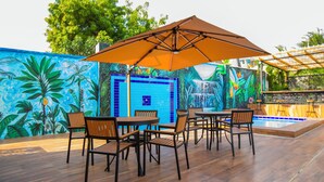 Eine Terrasse im Freien mit Holzmöbeln und einem großen Sonnenschirm, umgeben von farbenfrohen tropischen Wandgemälden.