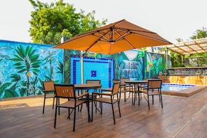 Un patio al aire libre con muebles de madera y un paraguas grande, rodeado de murales tropicales coloridos.
