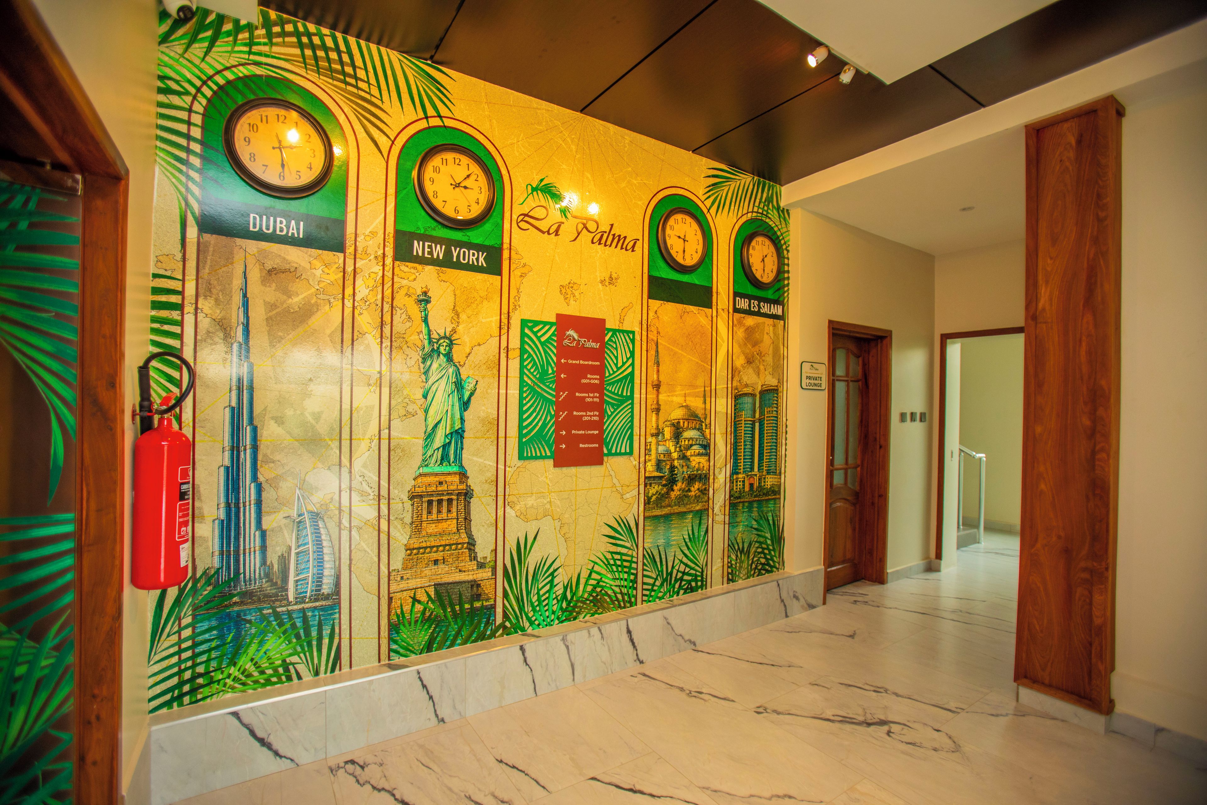 A lobby with a wall decorated with clocks and city landmarks, a fire extinguisher, and a marble floor.