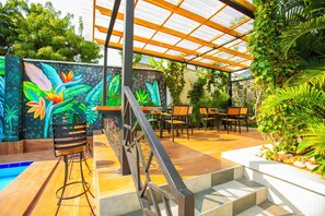 A covered outdoor seating area with a pool, surrounded by lush greenery and a vibrant mural.