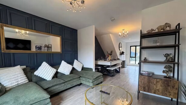 Ein modernes Wohnzimmer mit einem grünen Ecksofa, einem Glastisch und einem Essbereich, im Hintergrund ist eine Küche zu sehen.