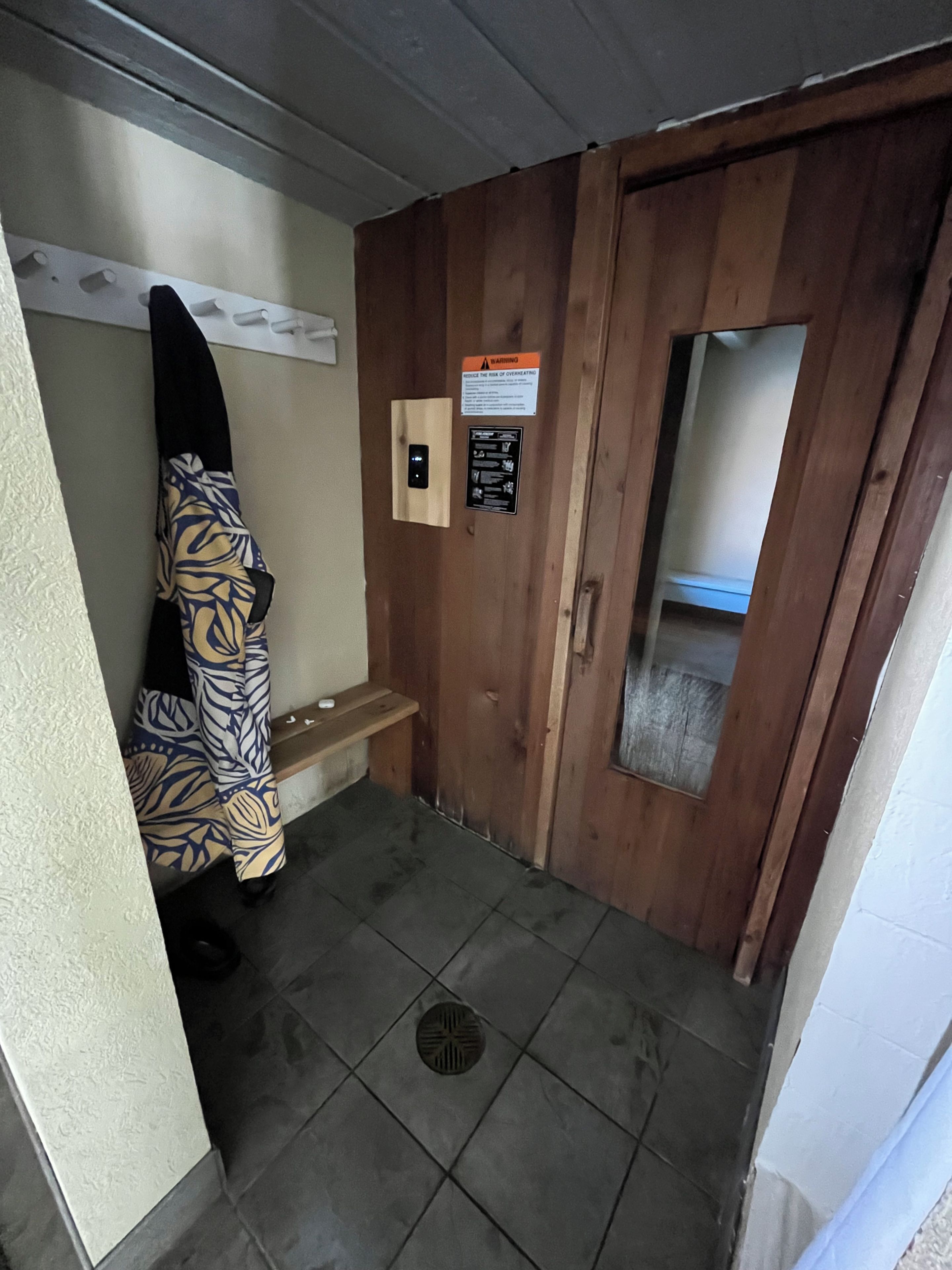 A small room with a wooden door, a bench, a towel rack, and a warning sign.