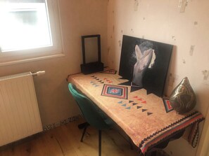 Et værelse med en radiator, en turkis stol, et bord med et mønstret borddække, et indrammet billede og en dekorativ vase.