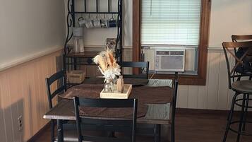 Un comedor con mesa y sillas de madera, un adorno temático de café en la pared y una ventana con persianas.