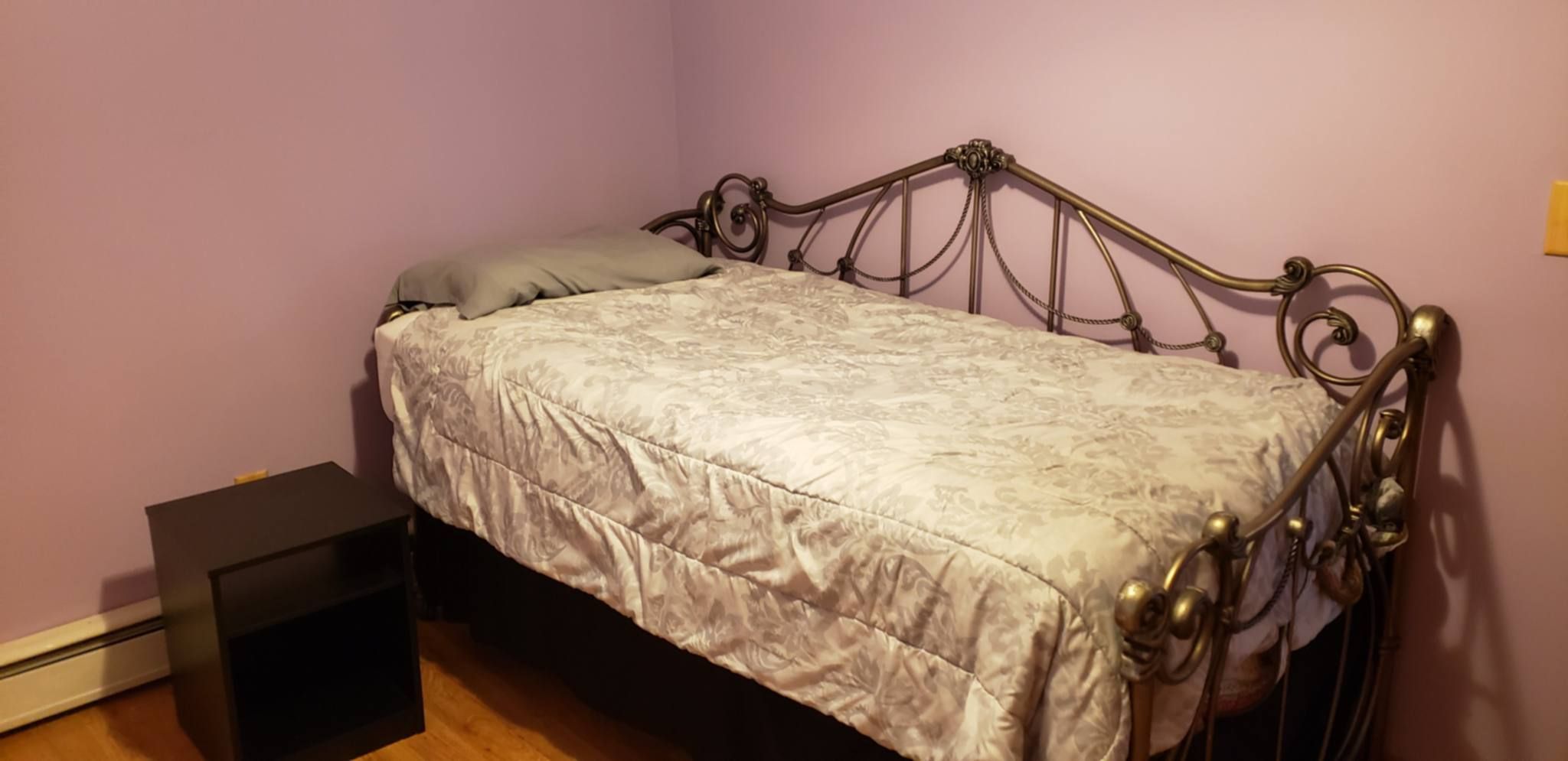 Una cama individual con estructura metálica y una colcha estampada en una habitación con piso de madera y una mesita de noche negra pequeña.
